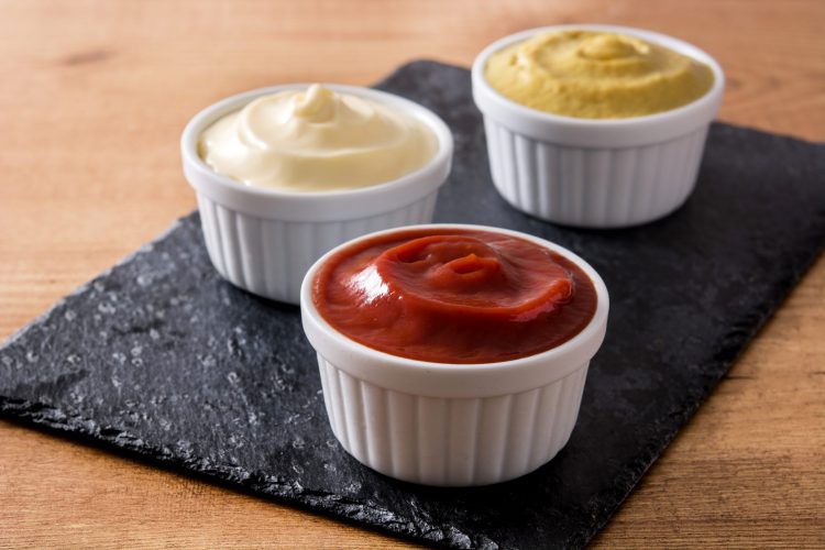 Ketchup, mosterd en mayonaise in witte kommetjes op een zwarte slat.
