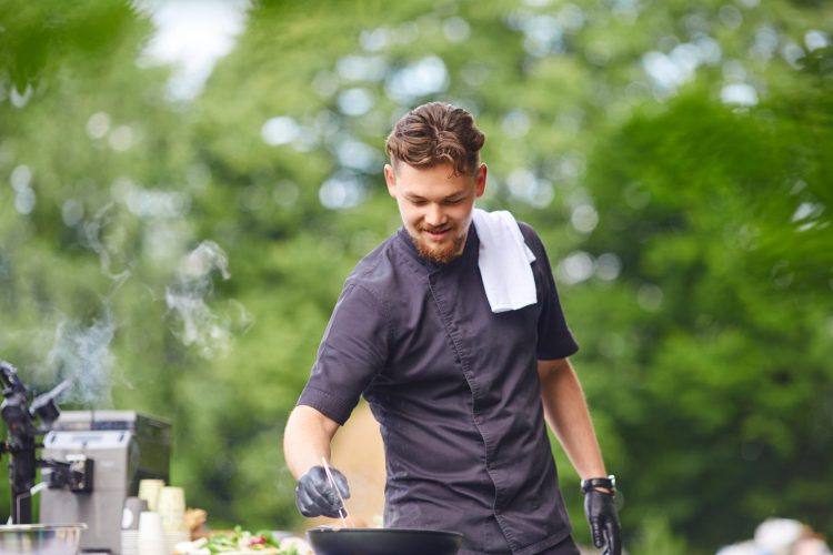 Een glimlachende knappe kaukasische mannelijke kok die zwarte outfit en witte handdoek op schouder in openlucht draagt.