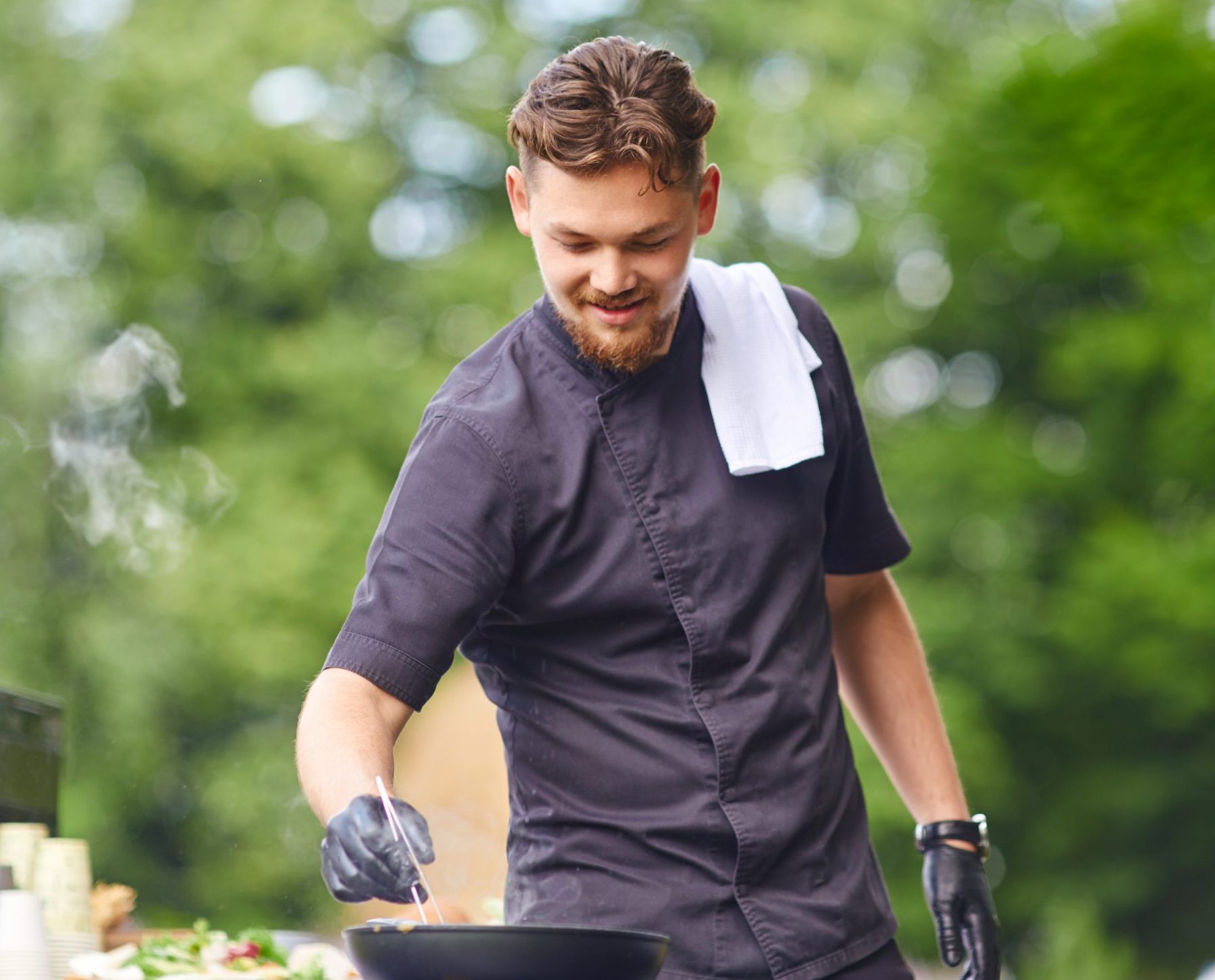Een glimlachende knappe kaukasische mannelijke kok die zwarte outfit en witte handdoek op schouder in openlucht draagt.