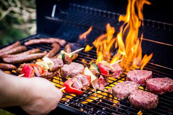 Gedetailleerde foto van rundvleesburgers, worsten en twee spiesjes op de barbecue waar ook vlammen bij te zien zijn.